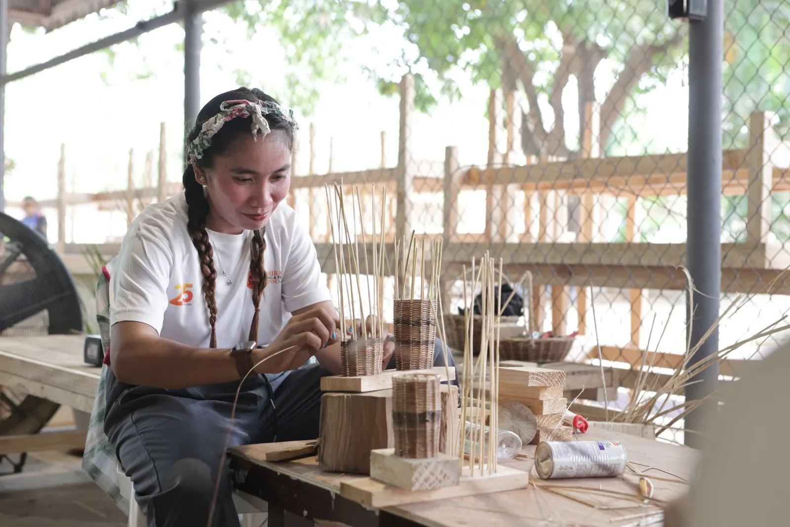 A woman wearing braids and a headband weaving a basket at a workshop table
